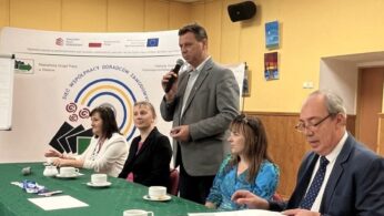 Pięć dorosłych osób siedzi przy zielonym stole z filiżankami i dokumentami w sali konferencyjnej. Jeden mężczyzna stoi i mówi do mikrofonu. Za nimi znajduje się baner z tekstem i logo oraz flipchart. Pokój ma żółte ściany i oprawione zdjęcia.