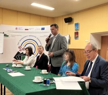 Pięć dorosłych osób siedzi przy zielonym stole z filiżankami i dokumentami w sali konferencyjnej. Jeden mężczyzna stoi i mówi do mikrofonu. Za nimi znajduje się baner z tekstem i logo oraz flipchart. Pokój ma żółte ściany i oprawione zdjęcia.
