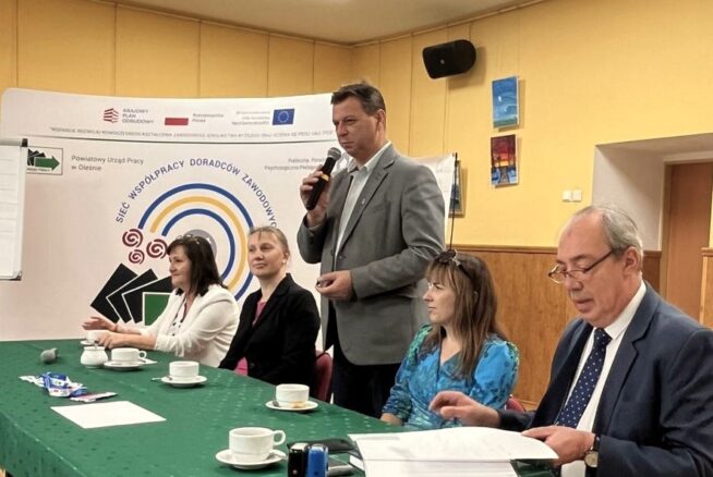 Pięć dorosłych osób siedzi przy zielonym stole z filiżankami i dokumentami w sali konferencyjnej. Jeden mężczyzna stoi i mówi do mikrofonu. Za nimi znajduje się baner z tekstem i logo oraz flipchart. Pokój ma żółte ściany i oprawione zdjęcia.