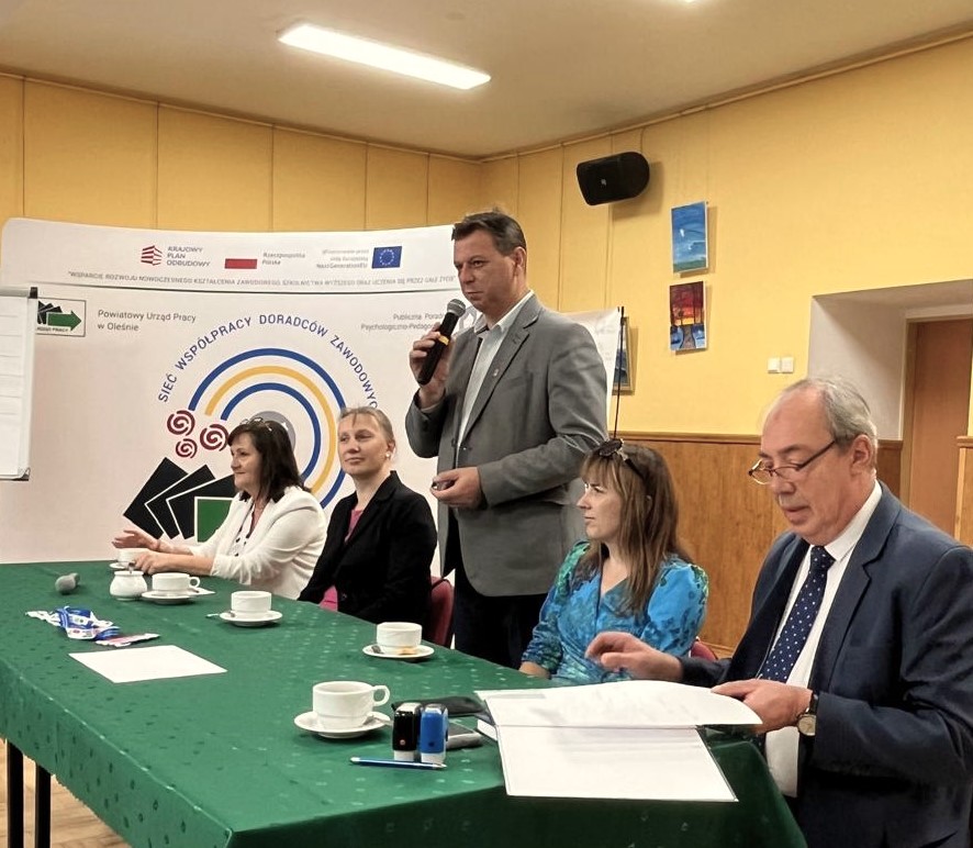 Pięć dorosłych osób siedzi przy zielonym stole z filiżankami i dokumentami w sali konferencyjnej. Jeden mężczyzna stoi i mówi do mikrofonu. Za nimi znajduje się baner z tekstem i logo oraz flipchart. Pokój ma żółte ściany i oprawione zdjęcia.