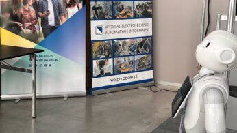 Biały humanoidalny robot stoi naprzeciwko dwóch banerów rekrutacyjnych Politechniki Opolskiej. Jeden baner przedstawia studentów i polski tekst o dołączeniu do uniwersytetu; drugi pokazuje zdjęcia z życia kampusu i wydziałów. Scenerią jest wewnętrzny korytarz.