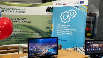 Na stole znajdują się dwa monitory komputerowe, z których jeden przedstawia krajobraz miasta z błyskawicami, a drugi interfejs połączenia wideo. Za nimi znajdują się dwa banery w języku polskim dotyczące szkoleń zawodowych i wsparcia zatrudnienia, z oficjalnymi logo i czerwonym balonem na lewej krawędzi.