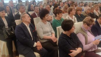 Duża grupa dorosłych w strojach formalnych i biznesowych siedzi w rzędach białych krzeseł w jasnym pomieszczeniu z dużymi oknami i kremowymi zasłonami, uważnie słuchając prezentacji lub mówcy poza kadrem.