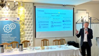 Mężczyzna w ciemnym garniturze mówi do mikrofonu na polskiej konferencji. Za nim ekran projekcyjny wyświetla Opolską pozytywną edukację 2021-2027. Po lewej stronie stoi niebieski baner finansowany przez UE. Na pierwszym planie długi stół z przekąskami.