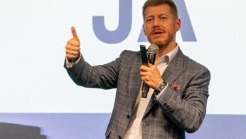 Mężczyzna z krótkimi jasnymi włosami i brodą, ubrany w kraciastą marynarkę, trzyma mikrofon w jednej ręce, a drugą podnosi kciuk do góry. Stoi przed ekranem wyświetlającym duże litery JA.