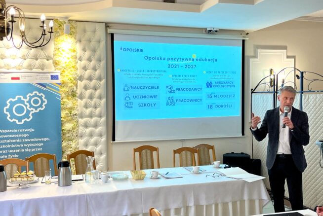 Mężczyzna w ciemnym garniturze mówi do mikrofonu na polskiej konferencji. Za nim ekran projekcyjny wyświetla Opolską pozytywną edukację 2021-2027. Po lewej stronie stoi niebieski baner finansowany przez UE. Na pierwszym planie długi stół z przekąskami.