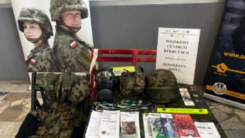 Stół z polskimi wojskowymi materiałami rekrutacyjnymi, mundurami maskującymi, hełmem, maskami przeciwgazowymi, broszurami i napisem 