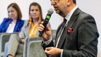 Mężczyzna w garniturze i okularach mówi do mikrofonu podczas wydarzenia. Dwie kobiety w strojach biznesowych siedzą w tle na szarych krzesłach, słuchając uważnie. Ostrość skupia się na mówcy, podczas gdy tło jest lekko rozmyte.