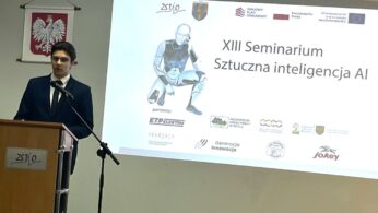 Młody mężczyzna w garniturze przemawia na podium z mikrofonem. Za nim znajduje się ekran projekcyjny wyświetlający 