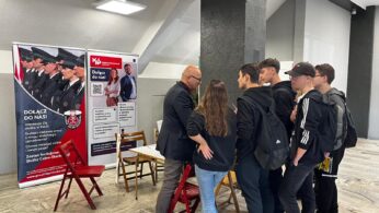Grupa młodych ludzi stoi przed stołem, rozmawiając z łysym mężczyzną w okularach na stoisku rekrutacyjnym. Za nimi znajdują się banery promujące karierę w policji i straży pożarnej, przedstawiające umundurowanych funkcjonariuszy i tekst informacyjny. Sceneria to wnętrze z szarymi ścianami.