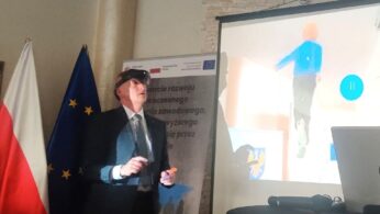Mężczyzna w garniturze i goglach VR stoi w pobliżu ekranu pokazującego wirtualną scenę spacerującej osoby. Za nim znajdują się flagi Polski, UE i regionalne oraz baner z polskim tekstem. Na stole na pierwszym planie znajduje się laptop.