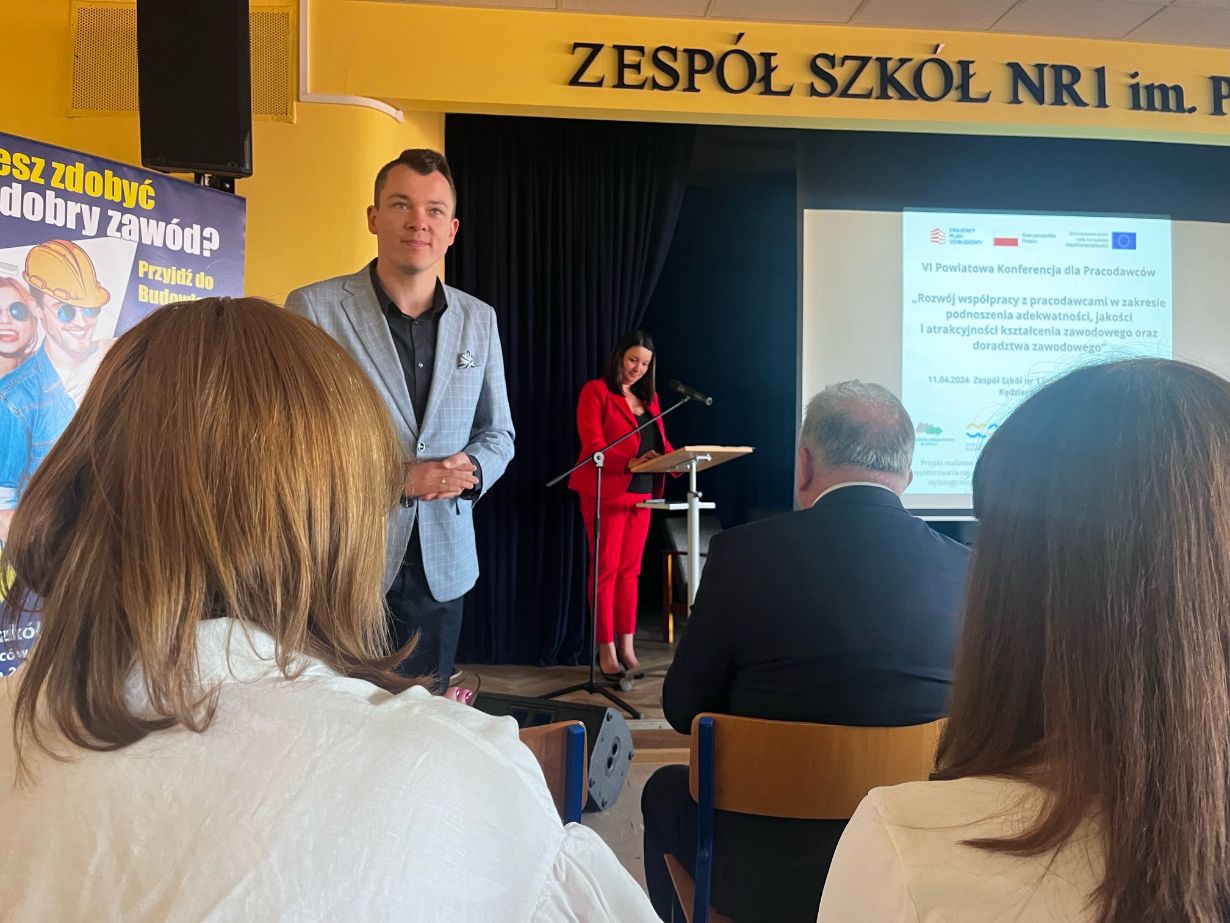 Mężczyzna w jasnoniebieskiej marynarce stoi na pierwszym planie, podczas gdy kobieta przemawia na podium na scenie. Kilka osób siedzi przodem do sceny. Wyświetlany jest slajd prezentacji, a nad nim widnieje żółty napis ZESPÓŁ SZKÓŁ NR1. Otoczenie wygląda na edukacyjne i formalne.