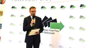 Mężczyzna w ciemnym garniturze i okularach trzyma mikrofon i teczkę, stojąc przed tłem ze strzałkami i polskim tekstem: Powiatowy Urząd Pracy w Głubczycach. Wygląda na to, że przemawia podczas oficjalnego wydarzenia.