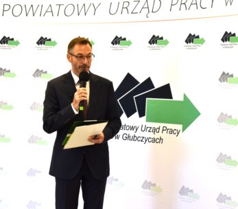 Mężczyzna w ciemnym garniturze i okularach trzyma mikrofon i teczkę, stojąc przed tłem ze strzałkami i polskim tekstem: Powiatowy Urząd Pracy w Głubczycach. Wygląda na to, że przemawia podczas oficjalnego wydarzenia.