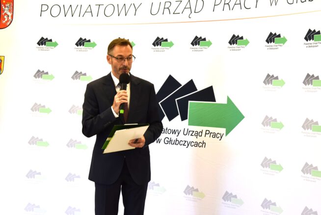 Mężczyzna w ciemnym garniturze i okularach trzyma mikrofon i teczkę, stojąc przed tłem ze strzałkami i polskim tekstem: Powiatowy Urząd Pracy w Głubczycach. Wygląda na to, że przemawia podczas oficjalnego wydarzenia.