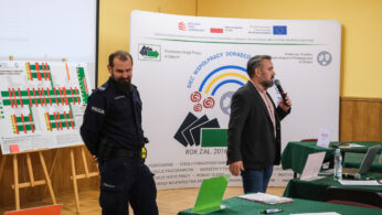 Mężczyzna w garniturze mówi do mikrofonu obok umundurowanego policjanta w klasie. Za nimi widoczna jest duża tablica prezentacyjna i wykresy. Zielone stoły z papierami i etykietami z nazwiskami wypełniają pomieszczenie, sugerując ustawienie spotkania lub warsztatu.