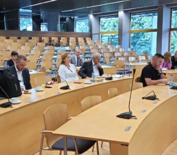 Grupa ludzi siedzi w nowoczesnej, jasnej sali wykładowej z dużymi oknami i drewnianymi meblami. Niektórzy uczestnicy są zwróceni do przodu, słuchając uważnie, podczas gdy inni patrzą w dół lub robią notatki. Sala jest w większości pusta, zajętych jest tylko kilka rzędów.