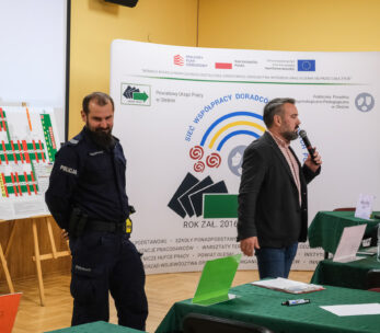 Mężczyzna w garniturze mówi do mikrofonu obok umundurowanego policjanta w klasie. Za nimi widoczna jest duża tablica prezentacyjna i wykresy. Zielone stoły z papierami i etykietami z nazwiskami wypełniają pomieszczenie, sugerując ustawienie spotkania lub warsztatu.