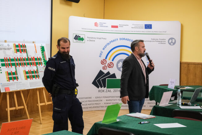 Mężczyzna w garniturze mówi do mikrofonu obok umundurowanego policjanta w klasie. Za nimi widoczna jest duża tablica prezentacyjna i wykresy. Zielone stoły z papierami i etykietami z nazwiskami wypełniają pomieszczenie, sugerując ustawienie spotkania lub warsztatu.