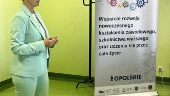 Kobieta w jasnoniebieskim garniturze, białej koszuli i trampkach stoi obok pionowego banera. Baner wyświetla polski tekst o wspieraniu nowoczesnej edukacji i uczenia się przez całe życie, z logo na górze i na dole. Pokój ma jasnozielone ściany i podłogę.