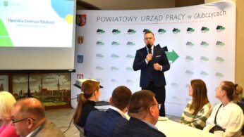 Mężczyzna w garniturze przemawia do mikrofonu na podium przed banerem z napisem Powiatowy Urząd Pracy w Głubczycach. Kilka siedzących osób słucha, niektórzy robią notatki. Wyświetlany slajd przedstawia Opolskie Centrum Edukacji i Ofertę dla rynku pracy.