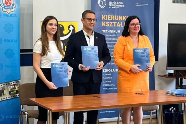 Trzy osoby stoją za stołem, trzymając niebieskie teczki i uśmiechając się do kamery. Są ubrani w formalne stroje. Za nimi znajdują się banery z polskim tekstem i logo, związane z policją i edukacją techniczną. Wygląda to na oficjalne wydarzenie lub ceremonię.