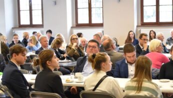Grupa ludzi, zarówno mężczyzn, jak i kobiet w różnym wieku, siedzi przy długich białych stołach w dobrze oświetlonym pomieszczeniu z dużymi oknami. Wydają się uważni i zaangażowani, niektórzy z filiżankami kawy i talerzami przed sobą, co sugeruje formalne spotkanie lub wydarzenie.