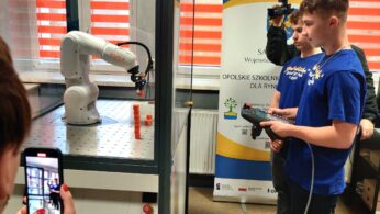 Dwóch nastolatków w niebieskich koszulkach obsługuje ramię robota za pomocą ręcznego kontrolera w laboratorium. Jedna osoba filmuje kamerą, a druga robi zdjęcie smartfonem. W tle widoczny jest baner i duże okna z czerwonymi roletami.