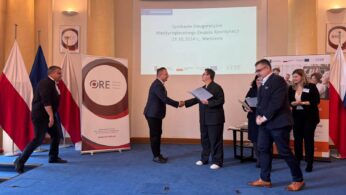 Sześć osób w oficjalnym pomieszczeniu z polskimi flagami i transparentami. Dwóch mężczyzn w garniturach podaje sobie ręce, trzymając dokumenty na środku sceny. Trzy osoby stoją po prawej stronie; jedna kobieta trzyma dokumenty, podczas gdy dwóch mężczyzn rozmawia. Jeden mężczyzna podchodzi do grupy z lewej strony.