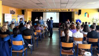 Grupa osób siedzi na niebieskich krzesłach naprzeciwko sceny w pomieszczeniu o żółtych ścianach. Z przodu stoi osoba prezentująca slajdy na ekranie. Niektórzy uczestnicy stoją lub poruszają się. Scena sugeruje wydarzenie lub spotkanie w szkole lub społeczności.