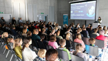Duża grupa ludzi, głównie dzieci, siedzi w rzędach krzeseł naprzeciwko mówcy na podium w nowoczesnej, jasnej sali konferencyjnej. Na dużym ekranie wyświetlana jest prezentacja zatytułowana KARIERA A CYFROWY SWIAT. Niektórzy dorośli stoją z tyłu w pobliżu drzwi wyjściowych.