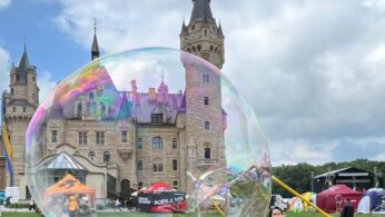 Dziewczynka w białym stroju tworzy gigantyczną bańkę mydlaną na trawiastym trawniku w pobliżu zamku z wysokimi iglicami. Inne dzieci i ludzie są w pobliżu, z kolorowymi namiotami i dmuchańcami w tle pod częściowo zachmurzonym niebem.