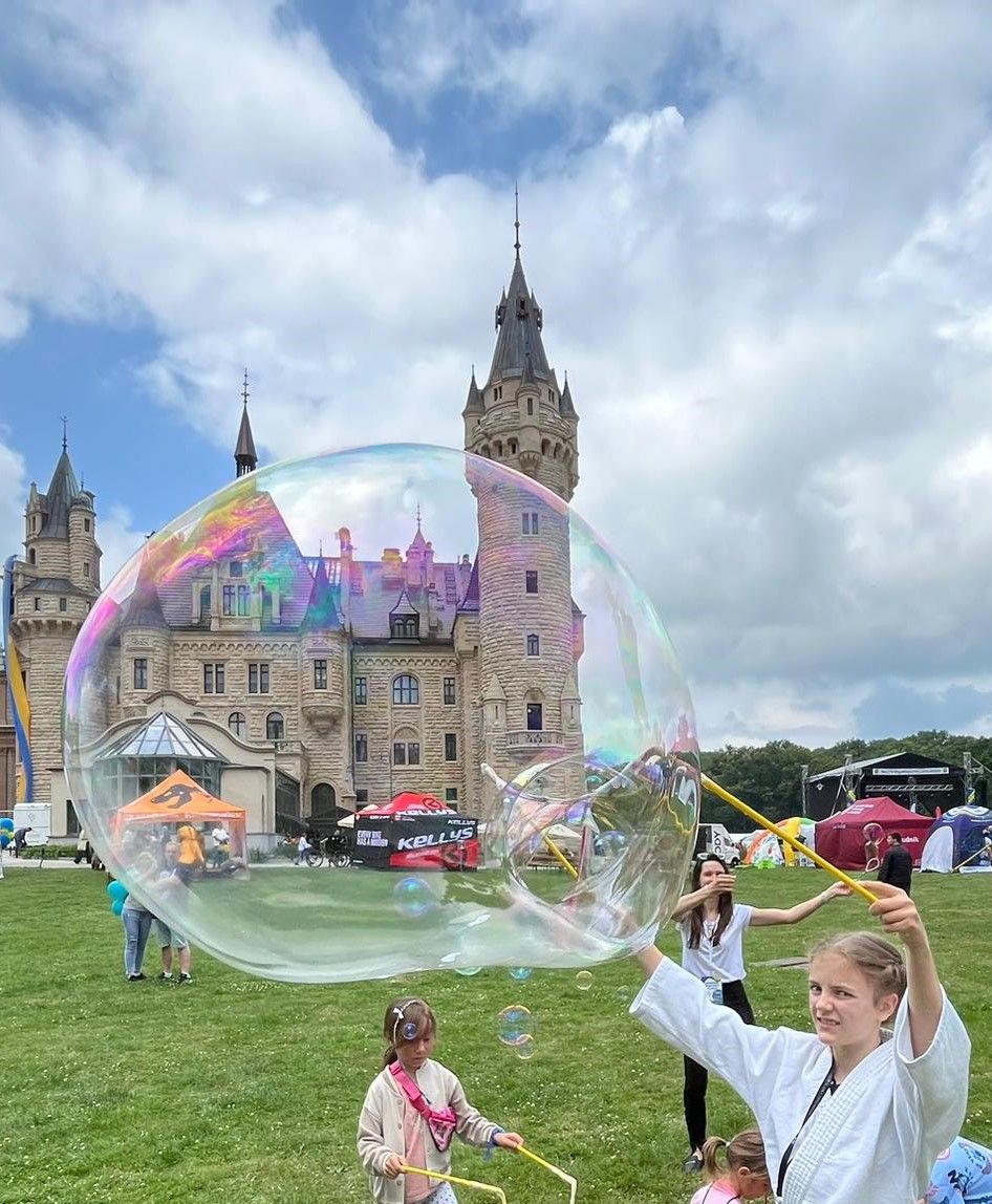 Dziewczynka w białym stroju tworzy gigantyczną bańkę mydlaną na trawiastym trawniku w pobliżu zamku z wysokimi iglicami. Inne dzieci i ludzie są w pobliżu, z kolorowymi namiotami i dmuchańcami w tle pod częściowo zachmurzonym niebem.