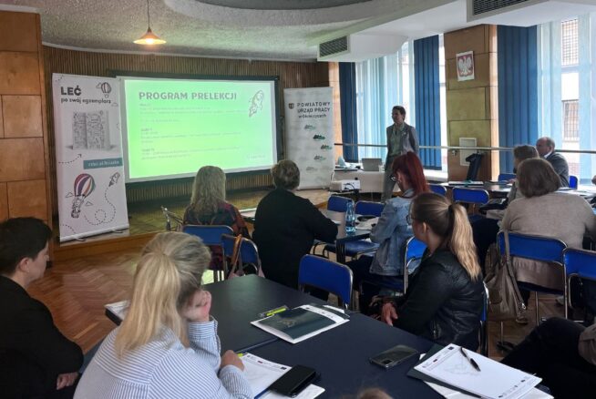 Prelegent stoi z przodu klasy i prowadzi prezentację. Uczestnicy siedzą przy biurkach naprzeciwko dużego ekranu wyświetlającego zielony slajd zatytułowany PROGRAM PRELEKCJI. Na ścianach wiszą plakaty i banery, a przez duże okna wpada światło dzienne.