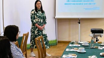 Kobieta w zielono-białej wzorzystej sukience stoi i przemawia przed wyświetlaną prezentacją w sali konferencyjnej. Siedzący uczestnicy słuchają przy stole z papierami, filiżankami kawy i przekąskami. Na slajdach i plakatach widnieją logotypy polskich i unijnych projektów.