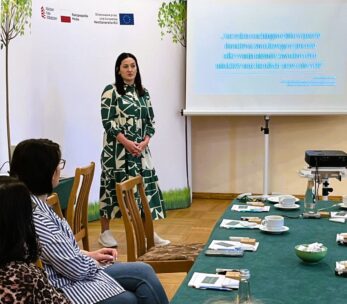 Kobieta w zielono-białej wzorzystej sukience stoi i przemawia przed wyświetlaną prezentacją w sali konferencyjnej. Siedzący uczestnicy słuchają przy stole z papierami, filiżankami kawy i przekąskami. Na slajdach i plakatach widnieją logotypy polskich i unijnych projektów.