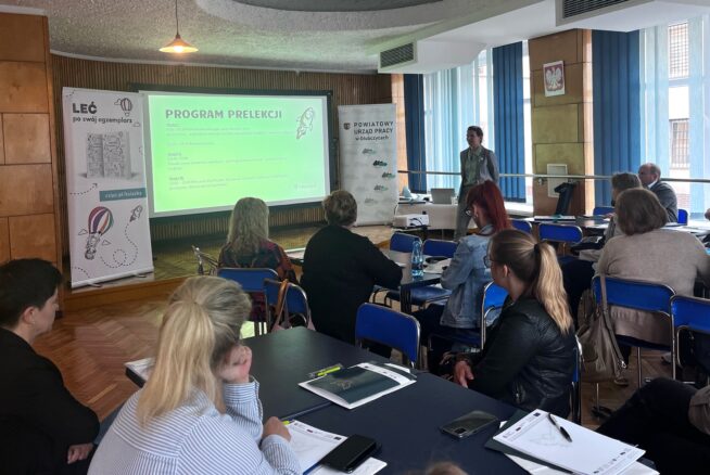 Grupa osób siedzi przy stołach w sali konferencyjnej, zwrócona przodem do mówcy. Na ekranie projektora wyświetlany jest zielony slajd zatytułowany PROGRAM PRELEKCJI. Niektórzy uczestnicy robią notatki. Pokój ma ściany wyłożone drewnianymi panelami i duże okna.