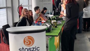 Grupa ludzi stoi i siedzi przy krytym stoisku wystawienniczym z banerami z napisem 