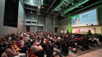 Duża publiczność siedzi w rzędach krzeseł naprzeciwko sceny w przestronnej hali w stylu industrialnym. Na scenie prezenterka w różowym stroju stoi w pobliżu ekranu wyświetlającego 