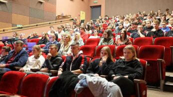 Duża grupa ludzi, głównie młodych dorosłych, siedzi w czerwonych fotelach teatralnych skierowanych do przodu w audytorium. Publiczność wydaje się być uważna, a niektórzy opierają głowy na dłoniach. Pokój ma beżowo-brązowe ściany i delikatne oświetlenie.