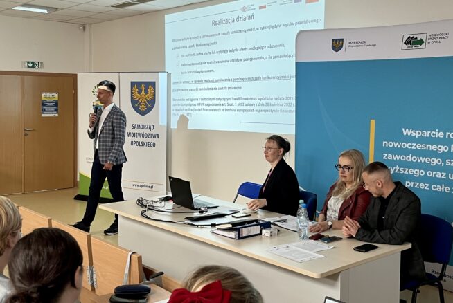 Mężczyzna stoi i mówi do mikrofonu obok ekranu projektora wyświetlającego polski tekst. Trzy osoby siedzą przy biurku z laptopami i dokumentami. Widoczne są banery z logo regionu i UE. Widzowie siedzą w rzędach na pierwszym planie.
