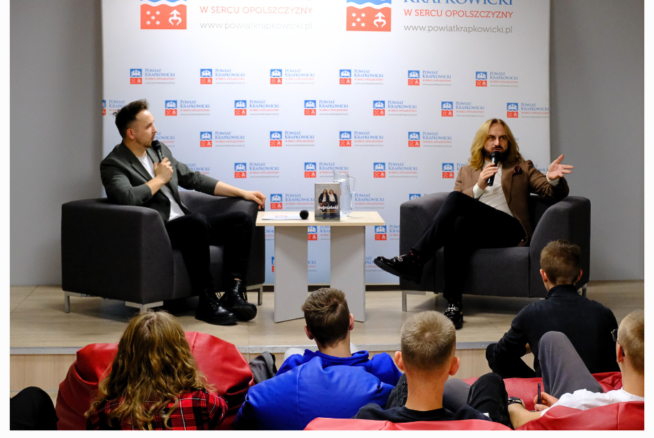 Dwóch mężczyzn siedzi w fotelach na scenie podczas dyskusji, jeden trzyma mikrofon i gestykuluje. Stoją twarzą do publiczności siedzącej na workach z fasolą. Za nimi znajduje się baner Powiatu Krapkowickiego z logo. Logotypy i flagi konferencji pojawiają się w dolnej części obrazu.