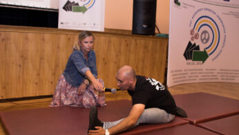 Kobieta klęczy na macie, wyciągając rękę, aby pomóc siedzącemu mężczyźnie wyciągnąć nogę do przodu. Mężczyzna trzyma mikrofon i dotyka swojej stopy. W tle wyświetlane są banery z logo i tekstem, a sceneria wydaje się być wydarzeniem społecznościowym w pomieszczeniu.
