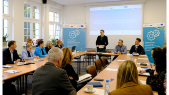 Grupa ludzi siedzi przy prostokątnych stołach w jasnej sali konferencyjnej. Kobieta stoi i przemawia w pobliżu dużego ekranu wyświetlającego prezentację. W tle widoczne są banery z kołami zębatymi i logo, a także okna.