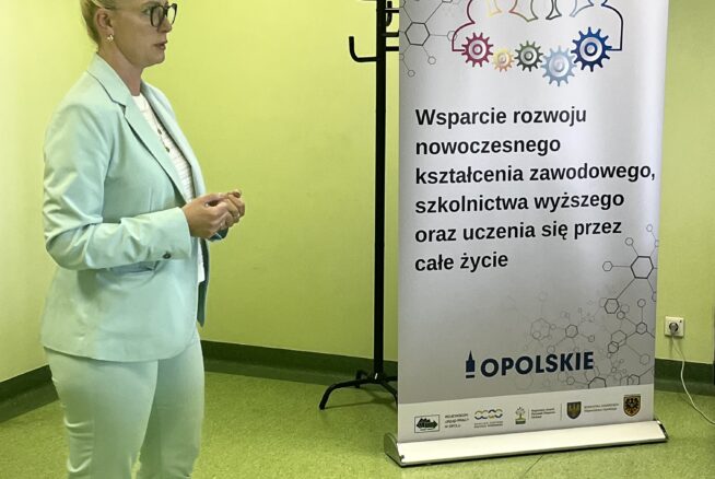 Kobieta w jasnoniebieskim garniturze, okularach i trampkach stoi w zielonym pokoju i przemawia. Obok niej znajduje się baner z polskim tekstem o wspieraniu nowoczesnego kształcenia zawodowego i uczenia się przez całe życie. Baner zawiera logo i abstrakcyjne postacie ludzkie.
