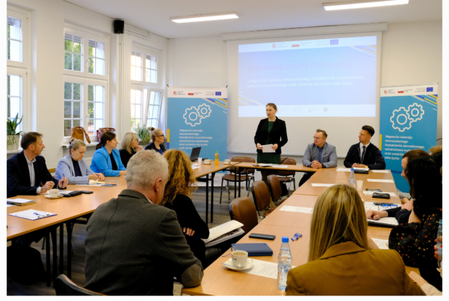 Grupa ludzi siedzi przy prostokątnych stołach w jasnej sali konferencyjnej. Kobieta stoi i przemawia w pobliżu dużego ekranu wyświetlającego prezentację. W tle widoczne są banery z kołami zębatymi i logo, a także okna.
