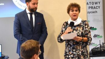 Kobieta w kwiecistej sukience mówi do mikrofonu, trzymając dokumenty. Obok niej stoi brodaty mężczyzna w granatowym garniturze. Trzeci mężczyzna siedzi na pierwszym planie. Za nimi częściowo widoczny jest baner i ekran projekcyjny. Otoczenie wydaje się formalne, prawdopodobnie konferencja.