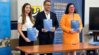 Trzy osoby stoją za drewnianym stołem, a każda z nich trzyma niebieską teczkę. Ubrani są w stroje biznesowe. Za nimi znajdują się banery z polskim tekstem i symbolami, w tym logo policji i godło szkoły. Po prawej stronie widoczny jest portret i ekran telewizora.
