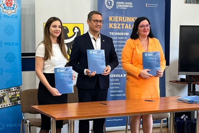 Trzy osoby stoją za drewnianym stołem, a każda z nich trzyma niebieską teczkę. Ubrani są w stroje biznesowe. Za nimi znajdują się banery z polskim tekstem i symbolami, w tym logo policji i godło szkoły. Po prawej stronie widoczny jest portret i ekran telewizora.