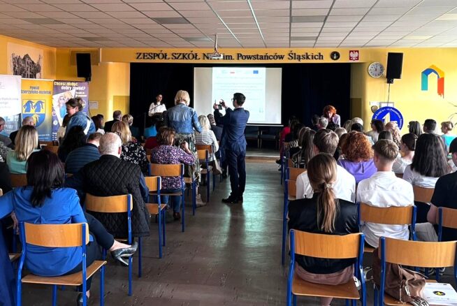 Grupa osób, w tym dorośli i uczniowie, siedzi naprzeciwko sceny w jasno oświetlonym pomieszczeniu o żółtych ścianach. Niektóre osoby stoją w pobliżu sceny, najwyraźniej prezentując. Duży ekran wyświetla prezentację z przodu pokoju.
